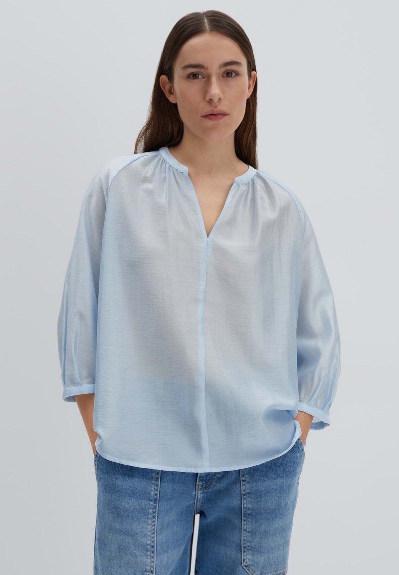 Blusa azzurro chiaro, semi-trasparente, con scollo a V, maniche arricciate e vestibilità ampia. Abbinata a pantaloni corti in denim, caratterizzati da una texture liscia.