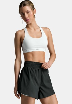 Donna atletica con reggiseno sportivo bianco e pantaloncini neri con fascia elastica in vita, in piedi davanti a uno sfondo semplice, con una mano appoggiata sulla vita dei pantaloncini.