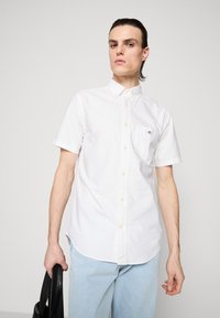 Egy fiatal férfi, hátrafésült sötét hajjal, fehér rövid ujjú gombos inget és világoskék farmernadrágot visel, bal kezében egy fekete táskát tart.