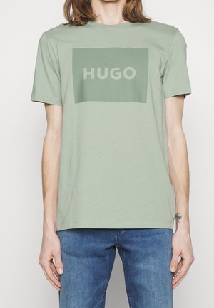 Lys grøn bomulds T-shirt med et rektangulært "HUGO" logo i en mørkere grøn. Korte ærmer og klassisk rund halsudskæring. Der bæres denim jeans.