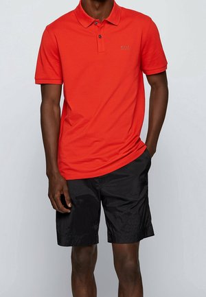 Rød polo i bomuld med krave, to knapper og broderet logo; kombineret med sorte shorts.