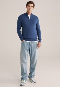 Pulover cu fermoar din material tricotat ușor de culoare navy, asortat cu blugi lejeri de un bleu deschis și adidași gri cu accente verzi.