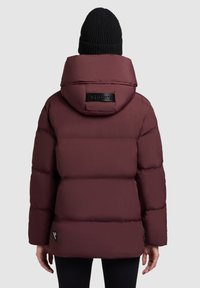 Bordeaux pufferjack met een capuchon, voorzien van een zwart logo patch op de achterkant, een gladde textuur en detail in de schoudernaden. Zwarte gebreide beanie gedragen.