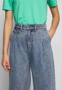 Vaaleansiniset korkeavyötäröiset ja leveälahkeiset farkut, joissa on kulunut pesu, etusovitukset, sivutaskut ja metallinen napitus.