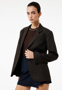 Brunternet blazer med en struktureret design, klokkerede lapeller og en enkelt knap. Båret over en mørk top og parret med en marineblå nederdel.