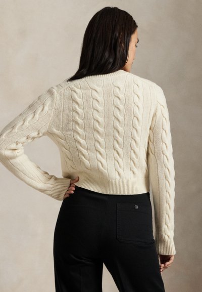 Vrouw met lang donker haar, in een crèmekleurige kabelgebreide trui en zwarte broek, staand met één hand in de zij, van achteren gezien.