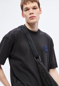 Μαύρο βαμβακερό μπλουζάκι με στρογγυλή λαιμόκοψη και κεντημένη μπλε κυκλική στάμπα· φορεμένο με υφασμάτινη, μαύρη τσάντα χιαστί με ιμάντα.