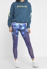 Felpa teal con dettagli giallo neon e cordino. Leggings aderenti con un motivo astratto blu e pannelli in rete trasparente. Sneakers bianche.