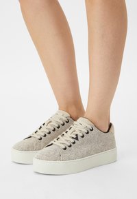 Baskets couleur crème avec une surface texturée, bout rond, œillets noirs et lacets beiges. Semelle blanche surélevée offrant une hauteur supplémentaire.