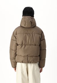 Brown puffer jacket with a high collar and hood, featuring horizontal quilting and a smooth exterior. Worn with light-colored pants.