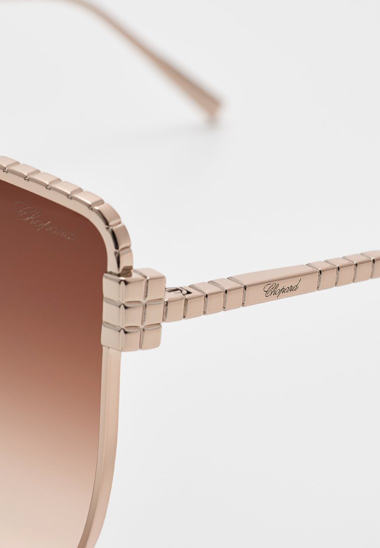 Grote zonnebril met een roségouden metalen frame, gebruinde gradient glazen, textuuraccenten op de armen en gegraveerd logo-detail.