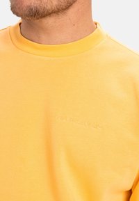 Sudadera de algodón amarilla con un cuello redondo acanalado. Presenta un logo bordado de "Quiksilver" en un tono más claro en el pecho.