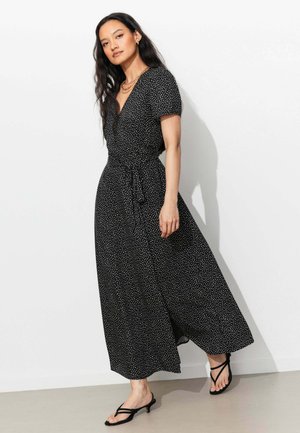 Femme aux longs cheveux foncés portant une robe midi noire à pois blancs et des sandales noires à brides avec talons kitten, debout contre un mur blanc.