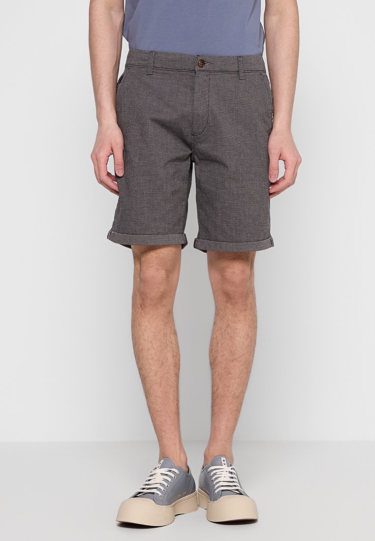 Homme portant un short gris à ourlet roulé, un t-shirt bleu et des baskets basses grises avec des lacets blancs, debout devant un fond uni.