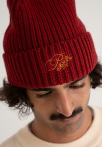 Červená pletená čepice s ribbovanou texturou a přeloženým lemem. Obsahuje vyšívané zlaté akcenty, včetně stylizovaných písmen a květinových vzorů.