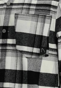 Chemise en flanelle à carreaux noirs et blancs avec une poche avant et des boutons-pression argentés, avec une petite étiquette noire avec un motif blanc.