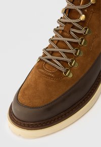 Braune Wildleder- und Lederwanderschuhe mit metallischen Ösen und beigen Schnürsenkeln. Sie verfügen über eine cremefarbene Gummisohle und genähte Akzente.