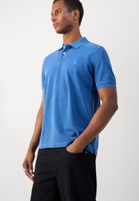 Blue cotton polo shirt with a collar, three-button placket, and small embroidered logo. Short sleeves, relaxed fit, paired with dark pants.