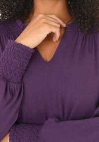V-kaulus violetti paita, jossa on rypytetyt hihat. Kangas näyttää pehmeältä ja teksturoidulta, ja siinä korostuu rypytetty yksityiskohta hihojen suutuksessa.