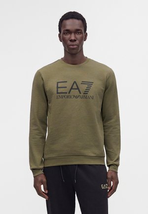 Homme portant un sweat-shirt vert olive avec un grand logo EA7 Emporio Armani et un pantalon noir avec un petit logo assorti sur la cuisse gauche.