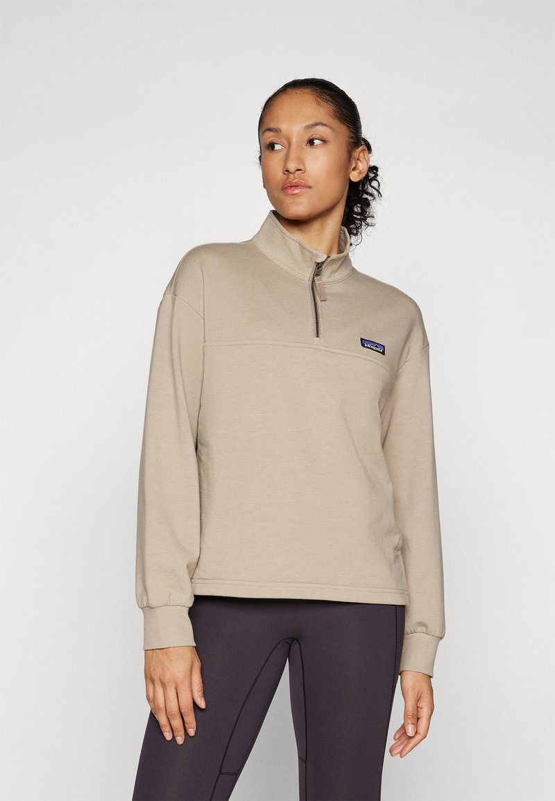 Beige kwart-rits pullover, zachte stof, lange mouwen, logo op de borst, relaxte pasvorm, gecombineerd met donkere legging, minimalistisch ontwerp.