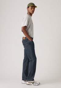 Lichtgrijze t-shirt met rode tekst, donkerblauwe jeans, groene pet en zilveren sportschoenen met zwarte accenten. Model staat in profiel.