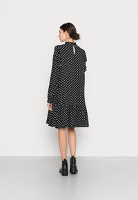 Vestido preto com bolinhas brancas, mangas longas, gola com volantes e barra com babados; apresenta um design de costas em forma de gota. Usado com botas pretas até o tornozelo.