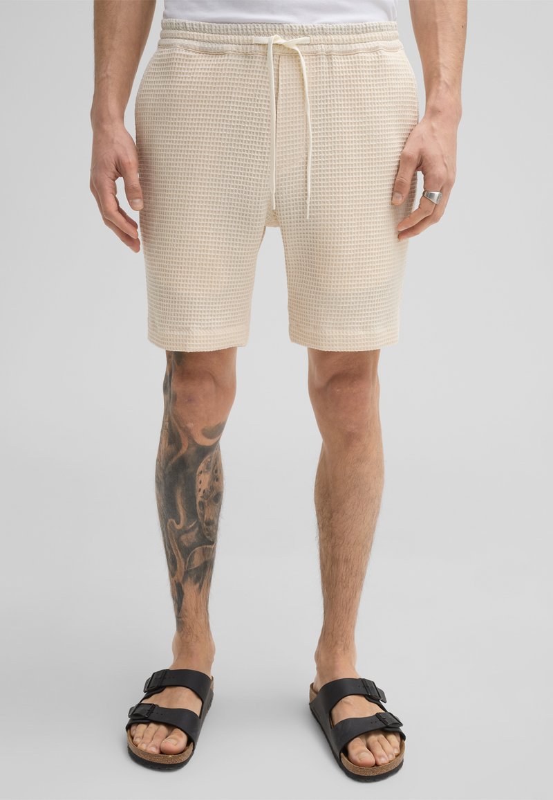 Beige Shorts mit Waffelstruktur, elastischem Bund und Kordelzug, ausgestattet mit zwei Seitentaschen und einem lockeren Schnitt. Schwarze Fußbekleidung sichtbar.