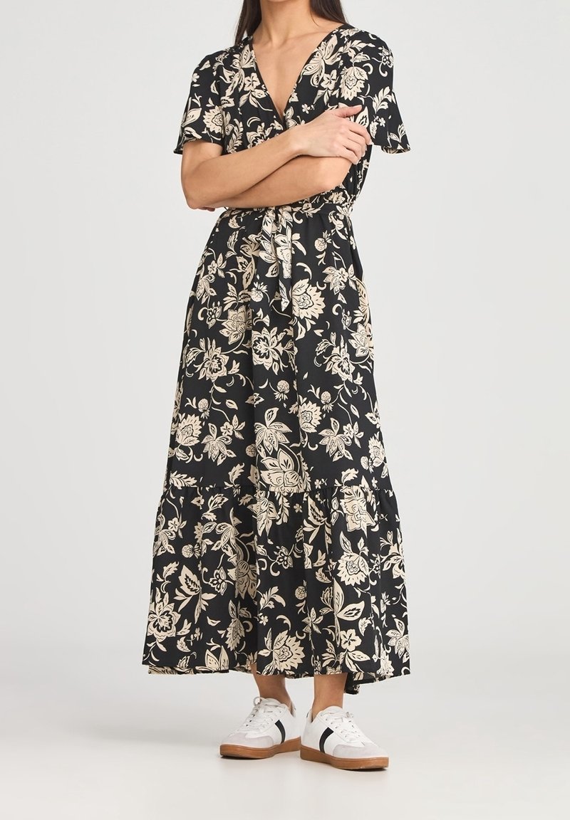 Femme portant une robe portefeuille noire à imprimé floral avec des manches courtes et des baskets blanches avec des rayures noires et des semelles marron.