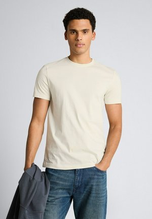 Młody mężczyzna z krótkimi kręconymi włosami, ubrany w beżowy t-shirt i niebieskie jeansy, trzymający ciemnoszarą kurtkę w jednej ręce.