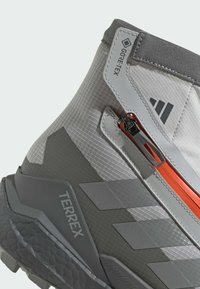 Grå vandringskänga med GORE-TEX-material. Har ett texturerat överdel, orange dragkedjedetalj och en robust gummisula med "TERREX"-logotypen.