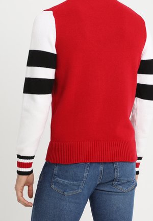 Muž v červeném svetr s bílými rukávy se dvěma černými pruhy a džínách, zobrazen zezadu na jednoduchém pozadí.