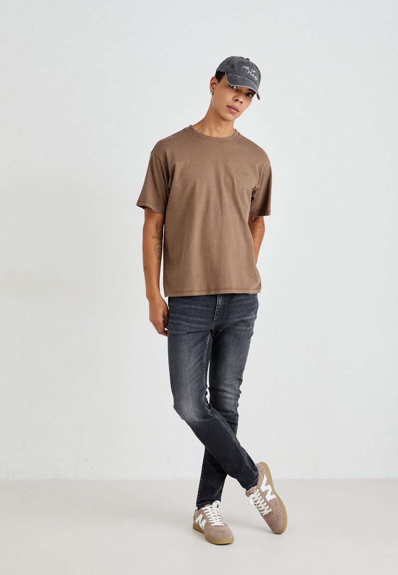 Braunes Baumwoll-T-Shirt, kurze Ärmel, Rundhalsausschnitt; dunkle Jeans; hellgraue Mütze mit Text; graue Sneakers mit weißen Akzenten.