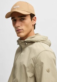 Beige cap with "BOSS" logo, worn with a light beige hooded jacket featuring a zip closure and textured fabric. Small logo on sleeve.