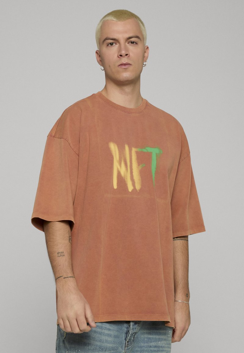 Oversized oranjebruin T-shirt van zachte stof, met een verloop "NFT"-afbeelding in geel en groen. Korte mouwen, ronde hals.