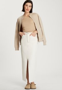 Beżowa kurtka bomberka, dopasowana brązowa bluzka, biała spódnica z denimu o wysokim stanie z rozcięciem z przodu, w połączeniu z brązowymi klapkami. Gładkie tekstury w całym stroju.