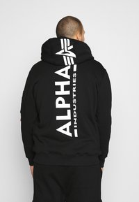 Felpa nera con cappuccio caratterizzata da un grande logo bianco "ALPHA INDUSTRIES" in verticale sulla schiena. Tessuto morbido con tasca a marsupio e cappuccio regolabile con cordino.