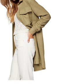 Femme portant un pantalon blanc, un haut en maille blanc et un long manteau vert olive avec une main dans la poche.