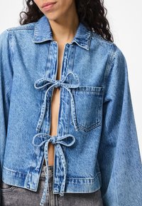 Lyseblå denimjakke med løs passform, knytninger foran og en brystlomme. Har krage og en lett slitt tekstur.