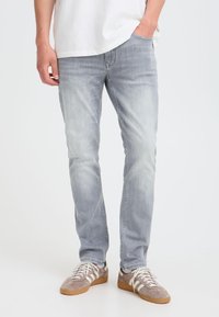Mand iført lysegrå slim-fit jeans og beige sneakers med hvide snørebånd, den ene hånd i lommen og hvid skjorte synlig ovenover.