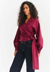 Bordowa bluzka kopertowa z kołnierzykiem, długimi bufkami i wiązaniem w pasie, zestawiona z ciemnymi dżinsami.