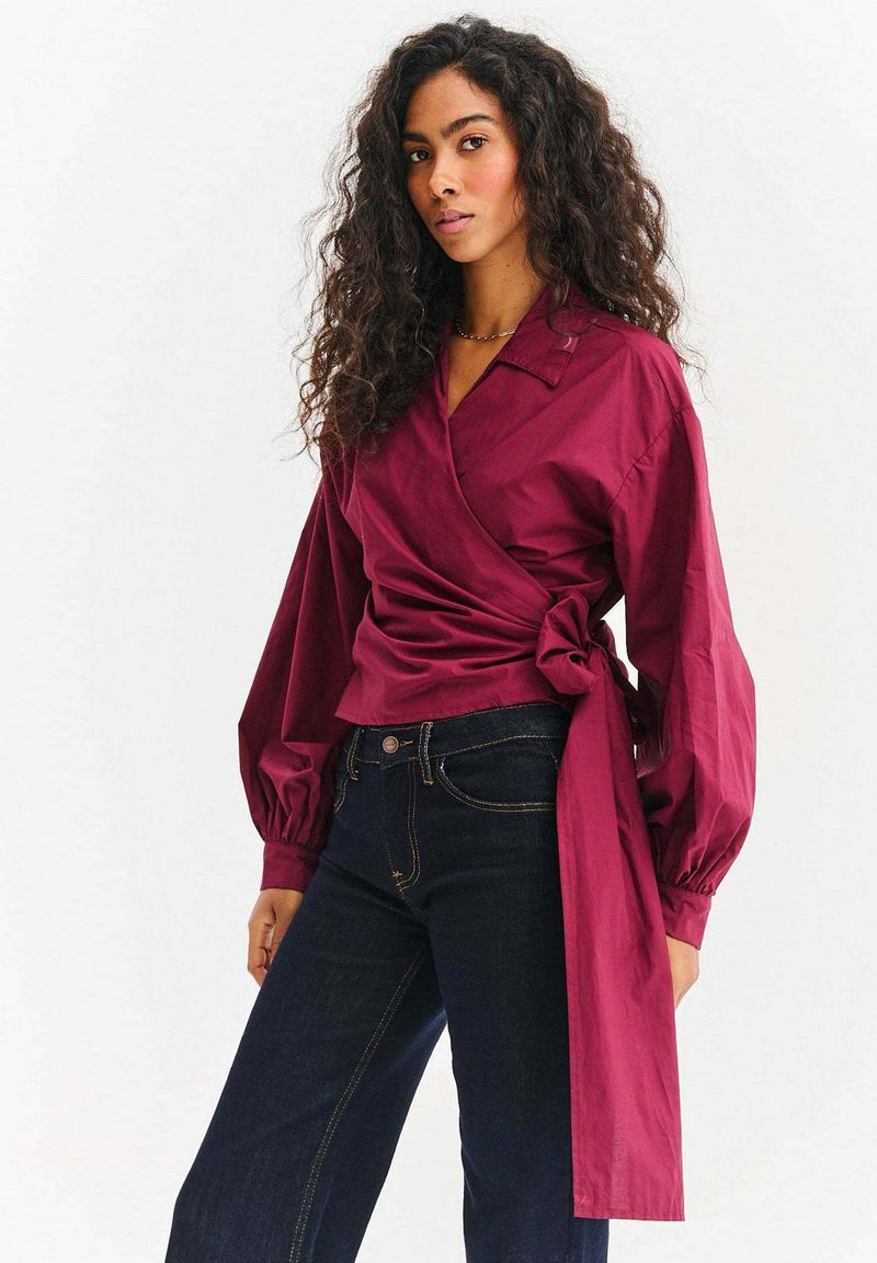 Bordowa bluzka kopertowa z kołnierzykiem, długimi bufkami i wiązaniem w pasie, zestawiona z ciemnymi dżinsami.