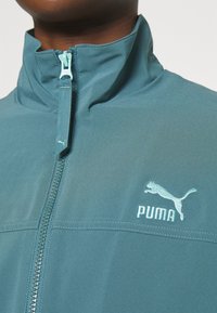 Teal Lynlåsjakke lavet af letvægtsmateriale med høj krave, PUMA-logo broderet i lyseblåt og en tekstureret lynlås.