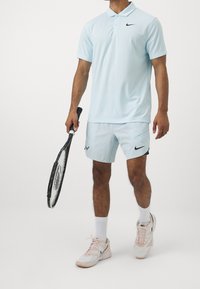 Jasnoniebieska koszulka polo z czarnym logo Nike, pasujące szorty, białe skarpetki i różowe tenisówki, trzymająca czarną rakietę tenisową.