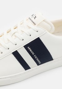 Sneaker bassa bianca con strisce blu navy e bianche. Presenta un tessuto texturizzato sulla linguetta e una patch con il marchio sul lato.
