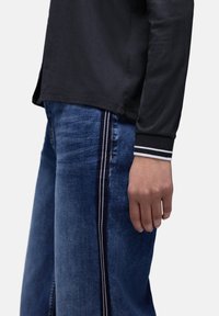 Chemise noire à manches longues avec un ourlet arrondi, associée à un jean bleu clair avec des bandes latérales blanches et bleu marine. Tissu lisse et coupe ajustée.