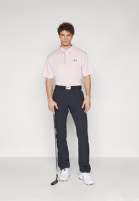Polo rosa de manga corta con cuello y logo negro, combinado con pantalones navy y un cinturón negro con hebilla plateada. Sostenido un palo de golf.