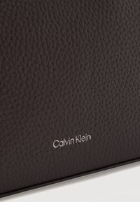 Υφή από σκούρο καφετί δέρμα με ανάγλυφο ασημένιο λογότυπο "Calvin Klein" κοντά στο κάτω άκρο.