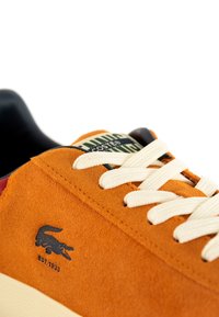 Sneaker in suede arancione con lacci crema, caratterizzata da un logo in cocco nero e un'etichetta della linguetta con motivo verde. Punta rotonda, design casual.