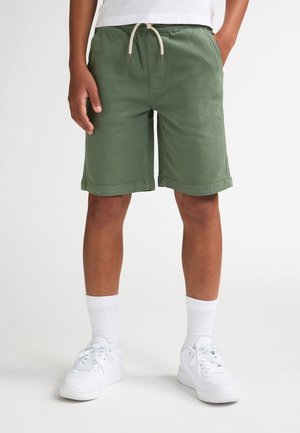 Groene katoenen korte broek met een elastische tailleband en trekkoord. Voorzien van zijzakken en een losse pasvorm. Draagt witte sokken en sneakers.